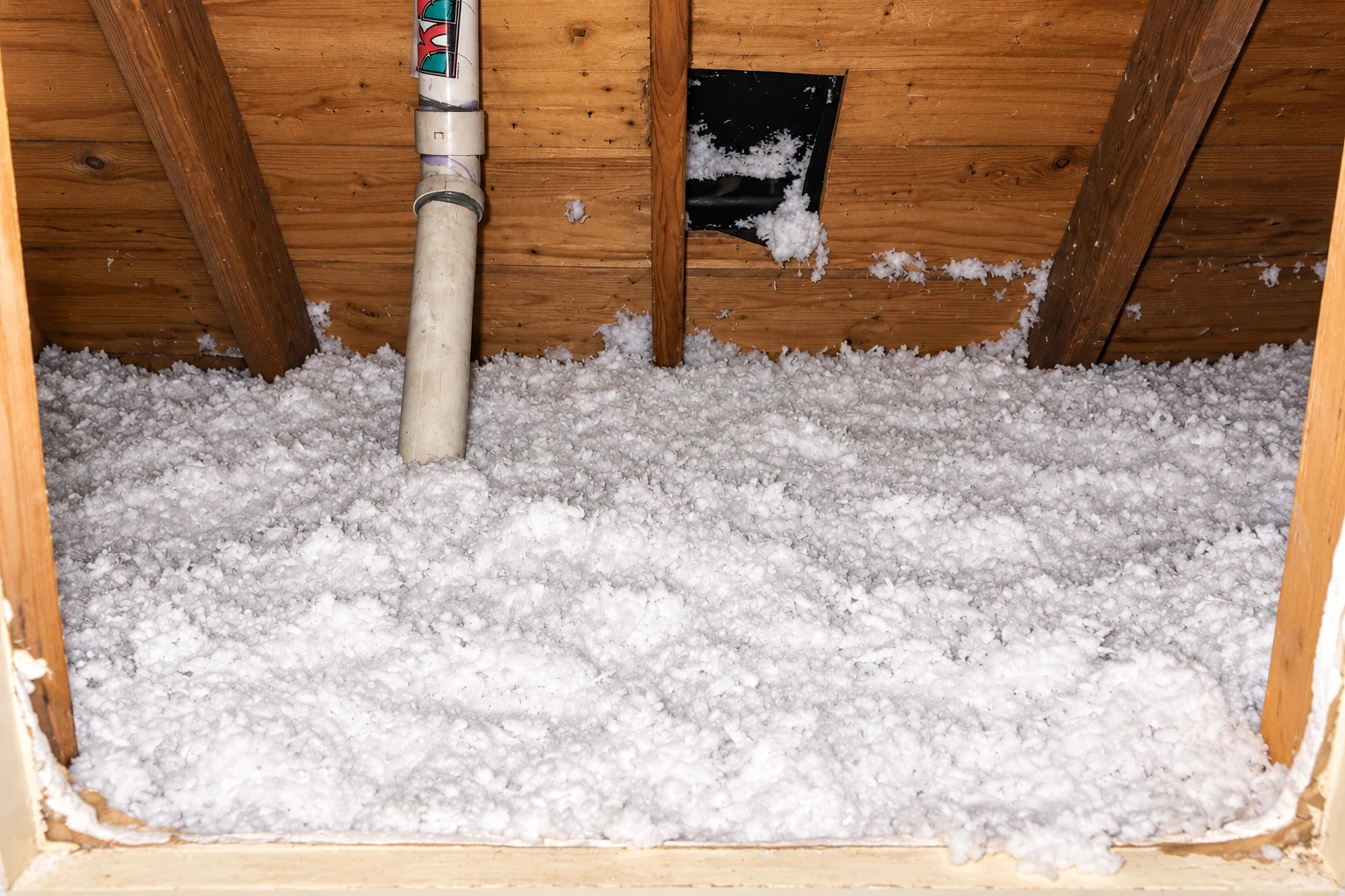 White blown-in insulation covers an attic floor beneath exposed wooden rafters and a vertical pipe.