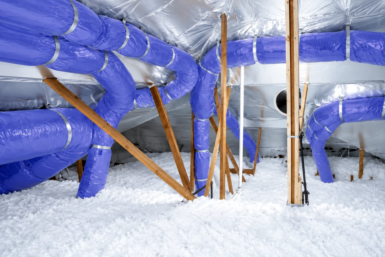 Blown-in insulation blankets an attic floor beneath wooden framing and a network of bright blue flexible ducts.
