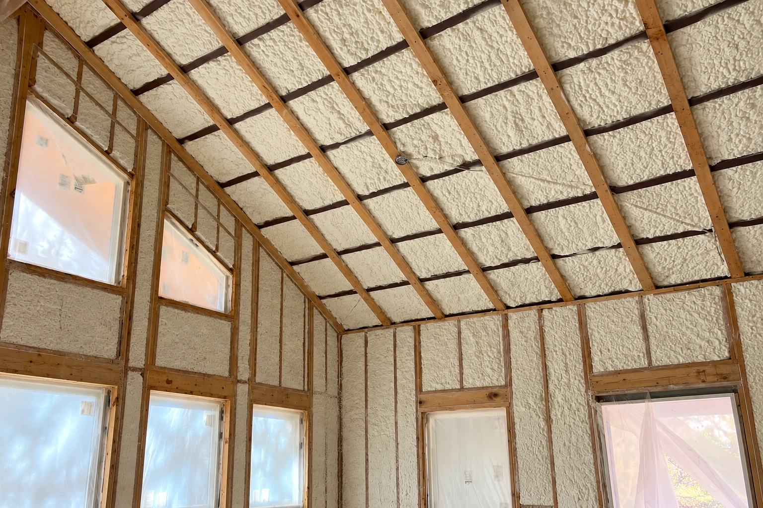 Sloped roof and walls of an unfinished room coated in cream-colored closed-cell spray foam around exposed wooden framing and windows.