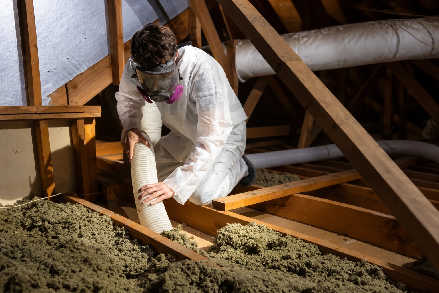 Worker in protective coveralls and a respirator uses a hose to remove loose attic insulation between wooden joists.
