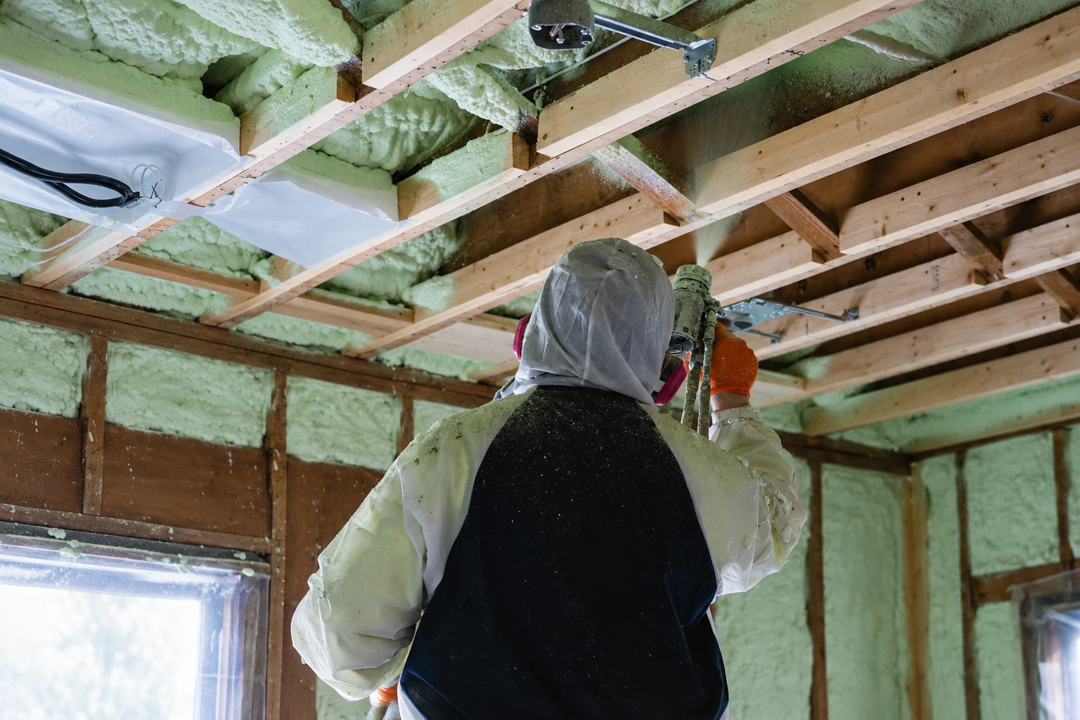 A worker in protective gear sprays pale green foam insulation between exposed ceiling joists in a wooden-framed room.