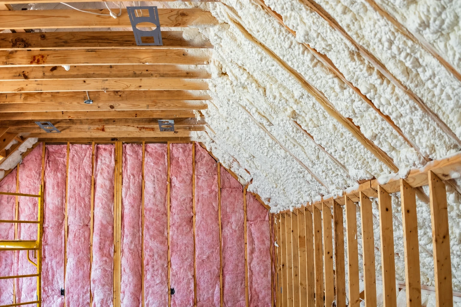 Spray foam insulation coats the sloped roof of a wood-framed attic, above wall cavities filled with pink batts.